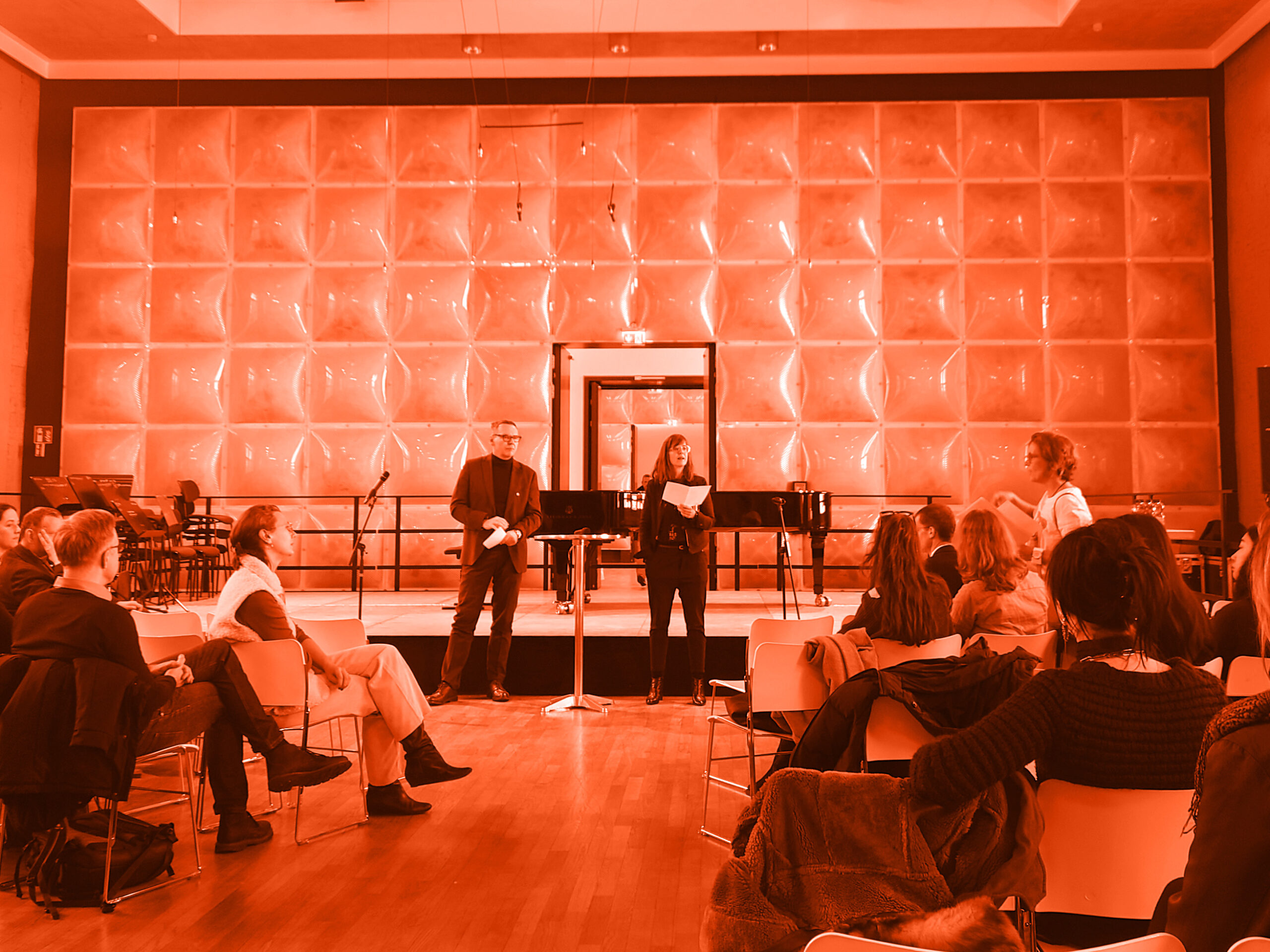 In einem modern gestalteten Saal sitzen Menschen auf Stühlen im Halbkreis und hören zwei Personen zu, die vorne stehen und sprechen. Im Hintergrund sind ein Flügel, Mikrofone und eine strukturierte Wand zu sehen; Das bild ist nachträglich orange gefärbt.