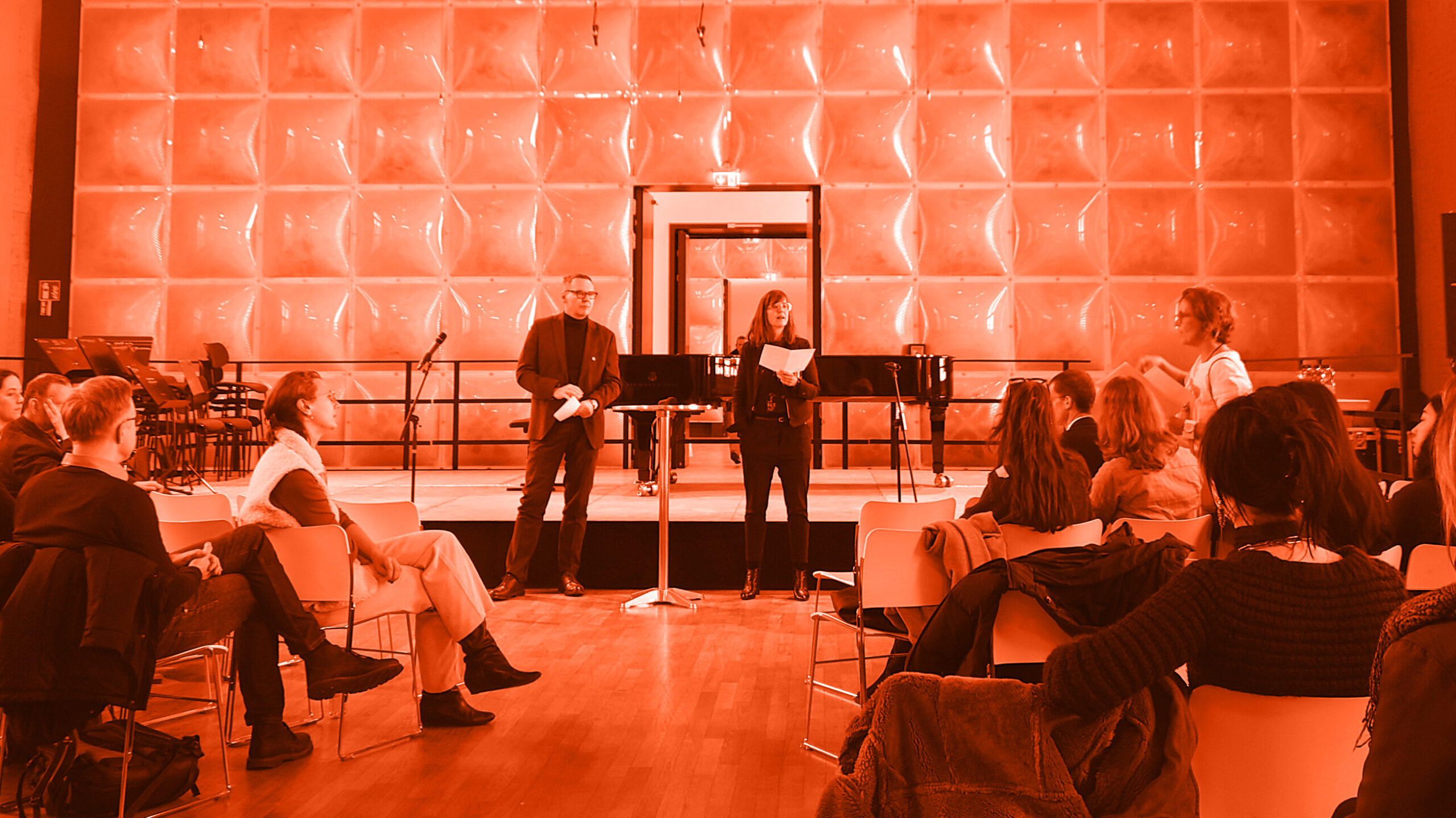In einem modern gestalteten Saal sitzen Menschen auf Stühlen im Halbkreis und hören zwei Personen zu, die vorne stehen und sprechen. Im Hintergrund sind ein Flügel, Mikrofone und eine strukturierte Wand zu sehen; Das bild ist nachträglich orange gefärbt.