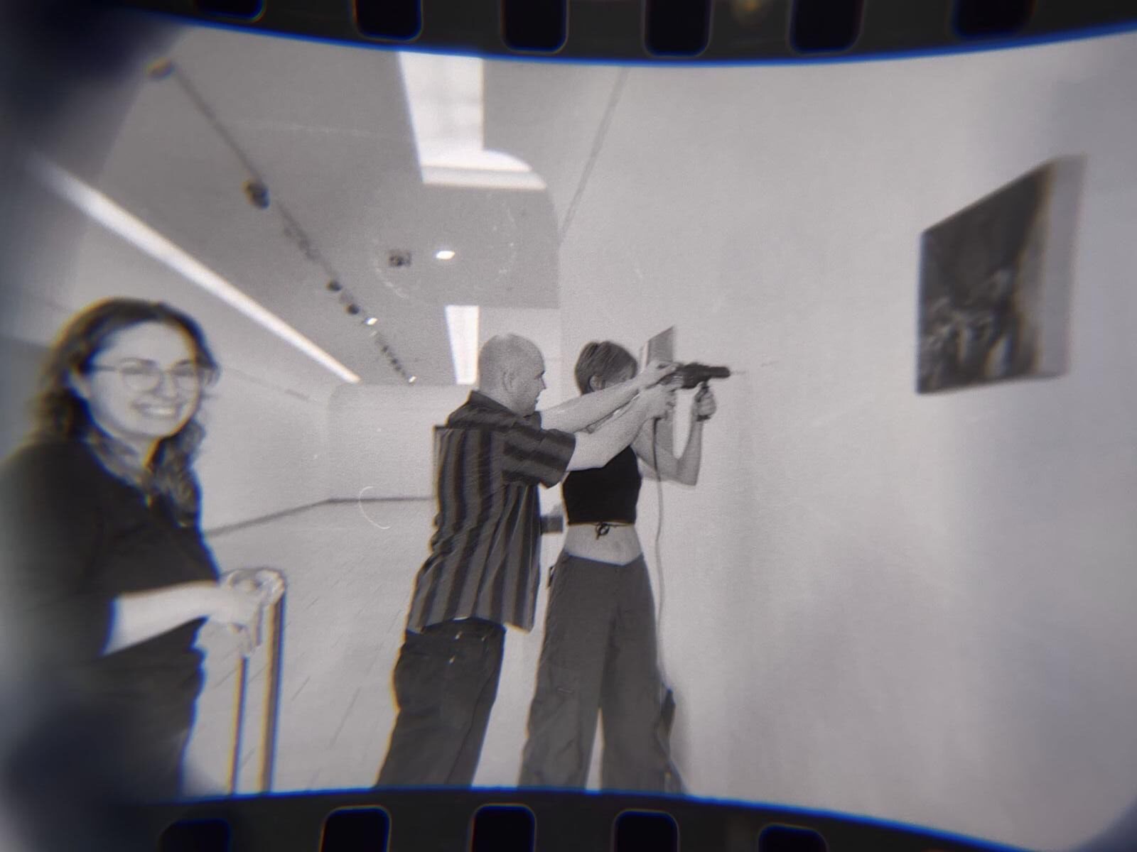 Blurred, analog, black-and-white photo with a visible film edge. It shows two person working on the wall with a drill, while an other person stands smiling on the left; the scene takes place in a bright interior with ceiling lights.