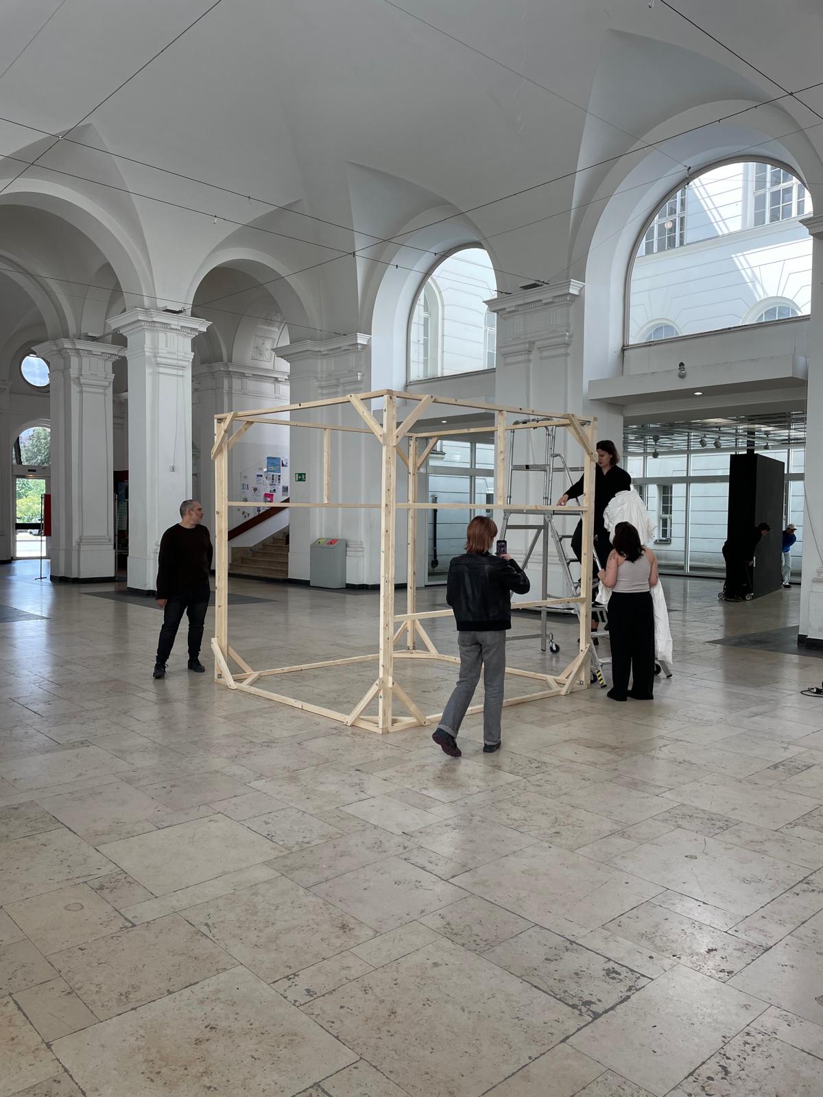 Five people assembling a large cube-shaped wooden frame in a bright, column-lined hall with arched windows (Foyer of Hardenbergstraße). One person is filming with a smartphone, another is standing on scaffolding working on the upper part of the frame.