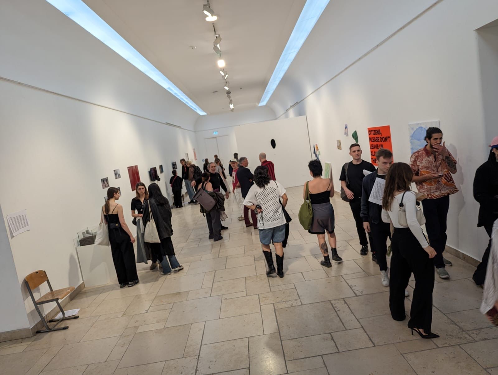 View into a long, brightly lit exhibition space with a light stone floor at Hardenbergstraße, where numerous visitors are gathered and viewing small-format artworks on the white walls.