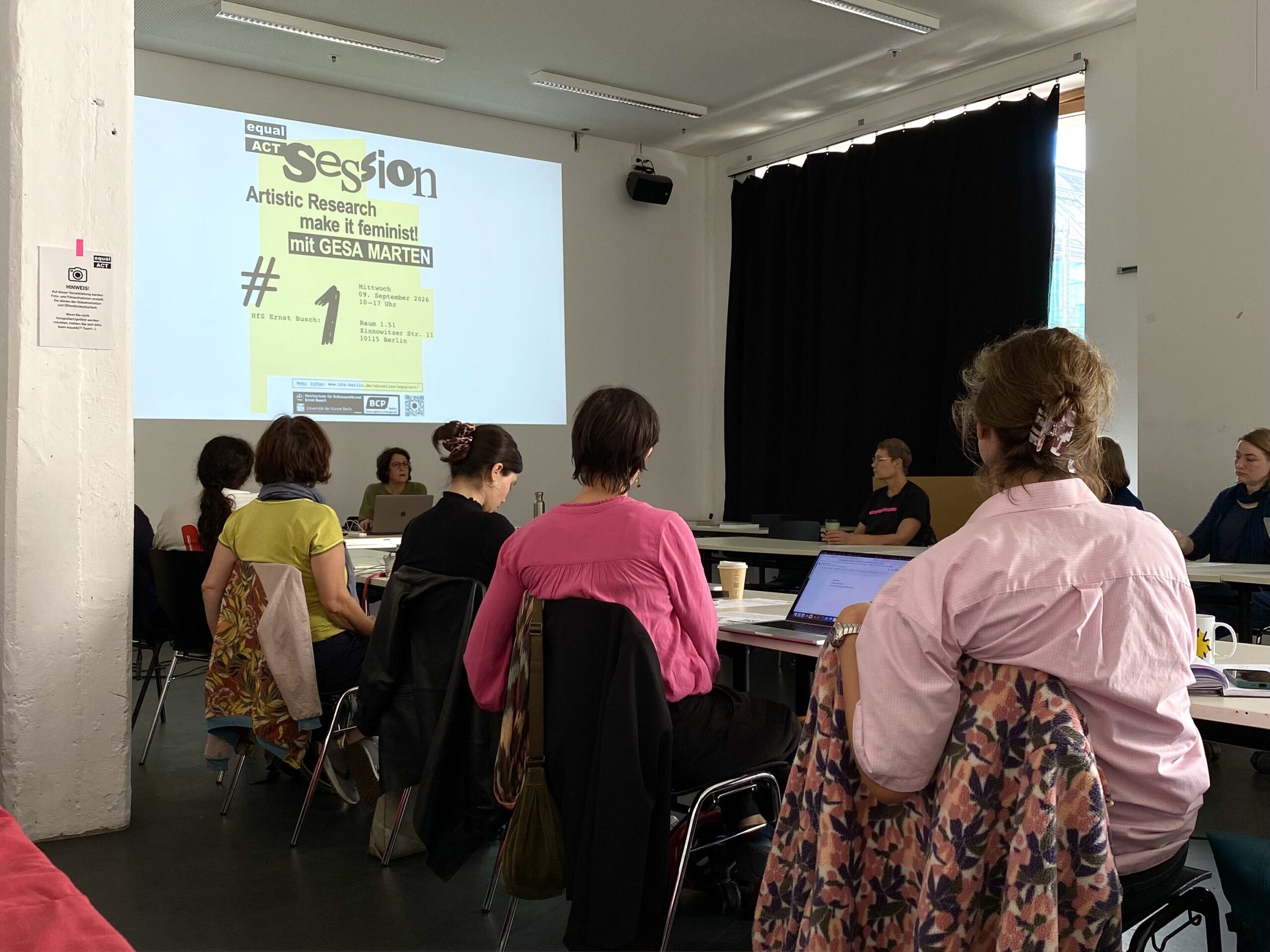 Teilnehmende einer Artistic-Research-Veranstaltung sitzen in einem Seminarraum der Hochschule für Schauspielkunst Ernst Busch. An der Wand ist das Plakat „Artistic Research – make it feminist!“ mit Gesa Marten projiziert. Die Teilnehmenden arbeiten an Tischen mit Laptops und Unterlagen.